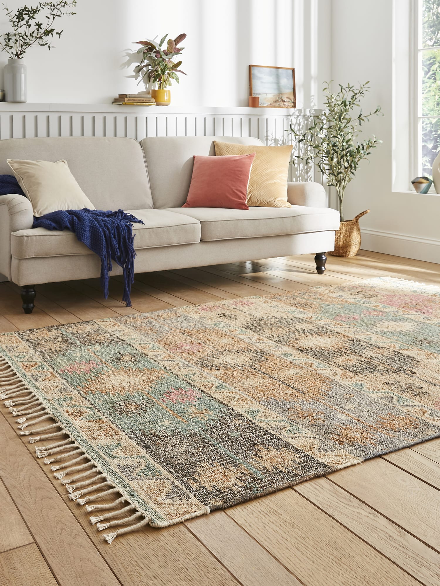 Bazaar Diamond Bands Multi Hand-Woven Rug. Bazaar Diamond Bands rectangular rug in mixed colours, at an angle showing the fringe beside a pale sofa.