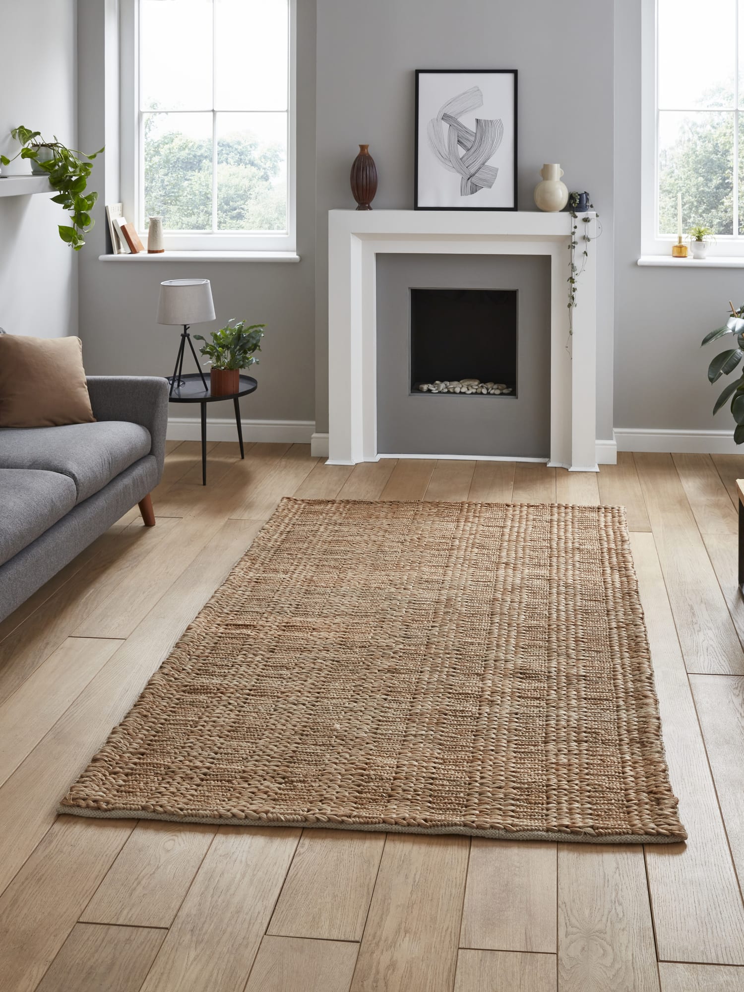 Bazaar Plain rectangular rug in natural, in front of a fireplace beside a grey sofa.