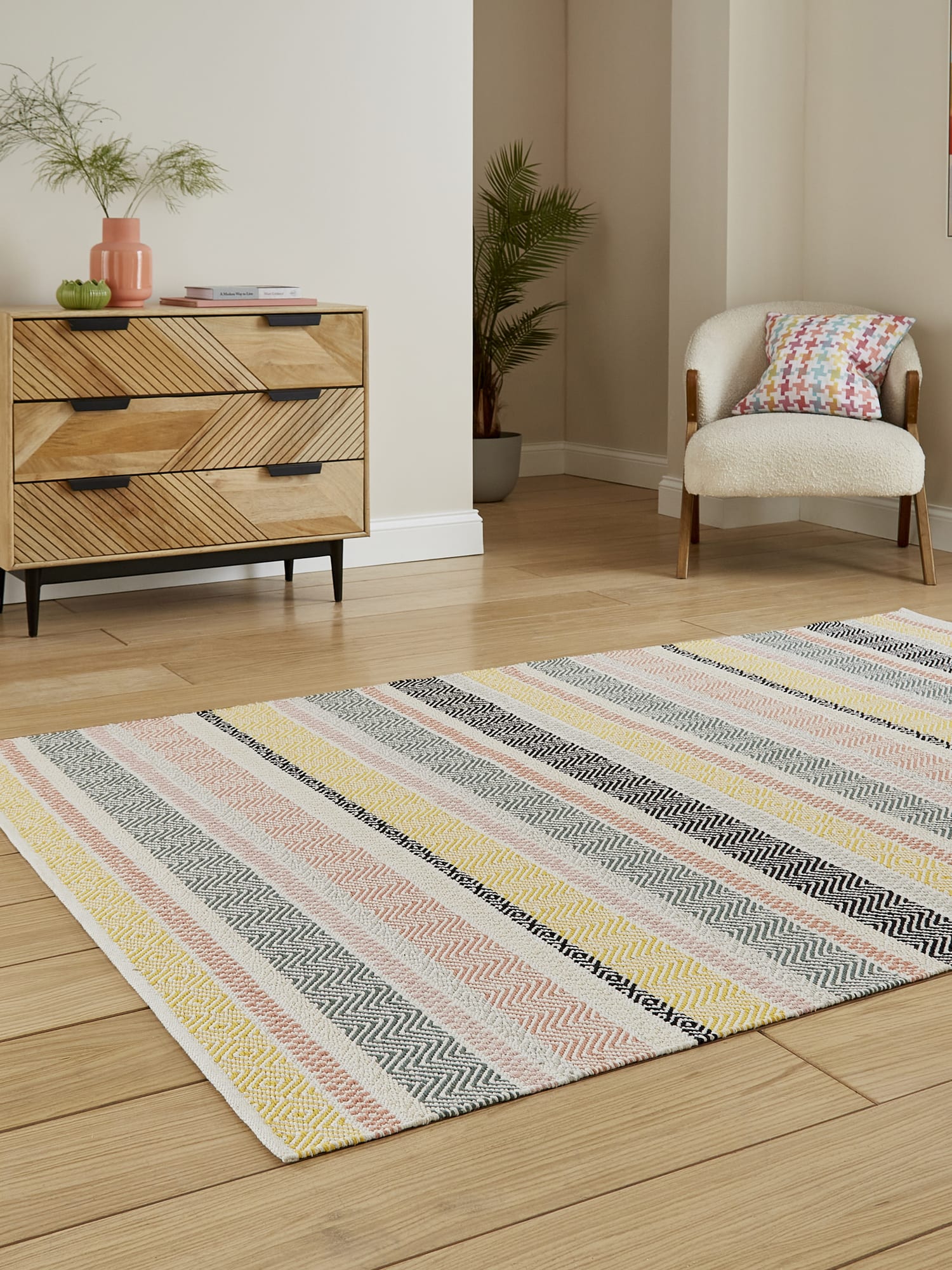 Cascade Pastel Multi Striped Cotton Rug. Cascade rectangular rug in pastel mixed colours, at an angle beside a wooden chest of drawers.