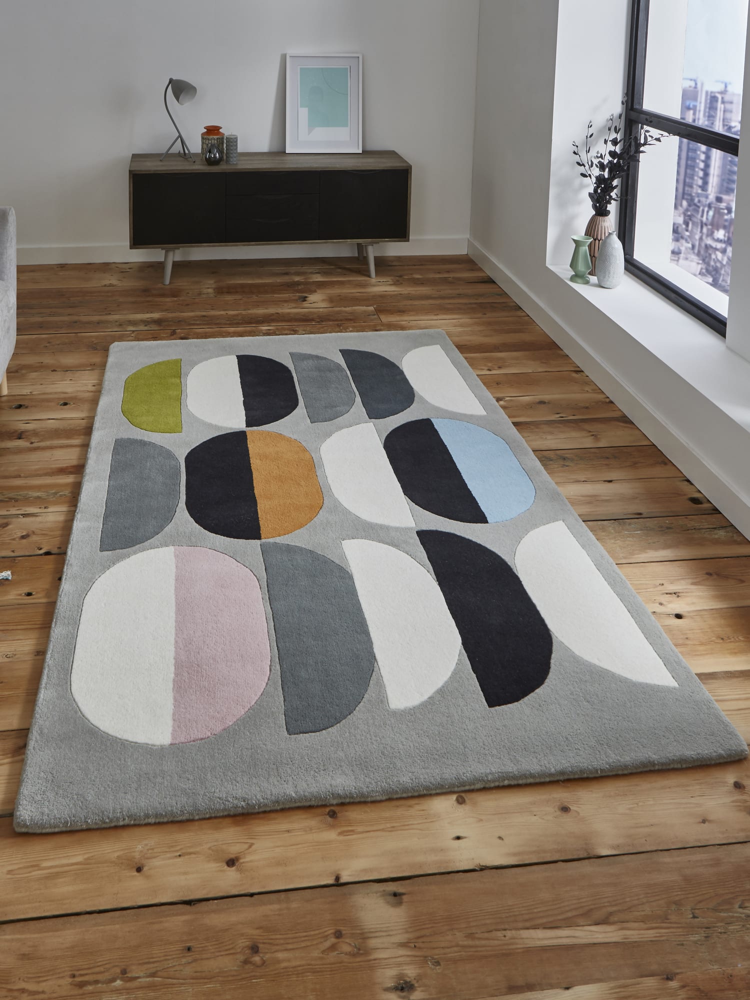 Inaluxe Composition rectangular rug in grey, black and cream with yellow, blue and pink accents, on a wooden floor beside a window.