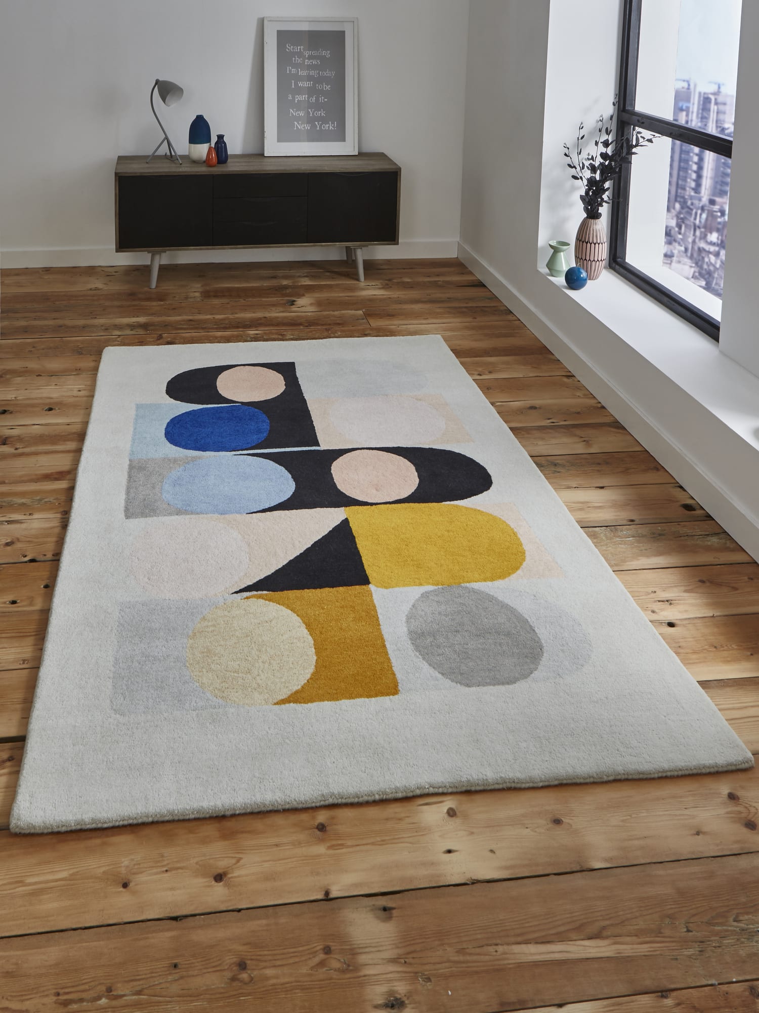 Inaluxe Jazz Flute rectangular rug in grey and cream with blue, yellow, pink and black shapes, on a wooden floor beside a window.