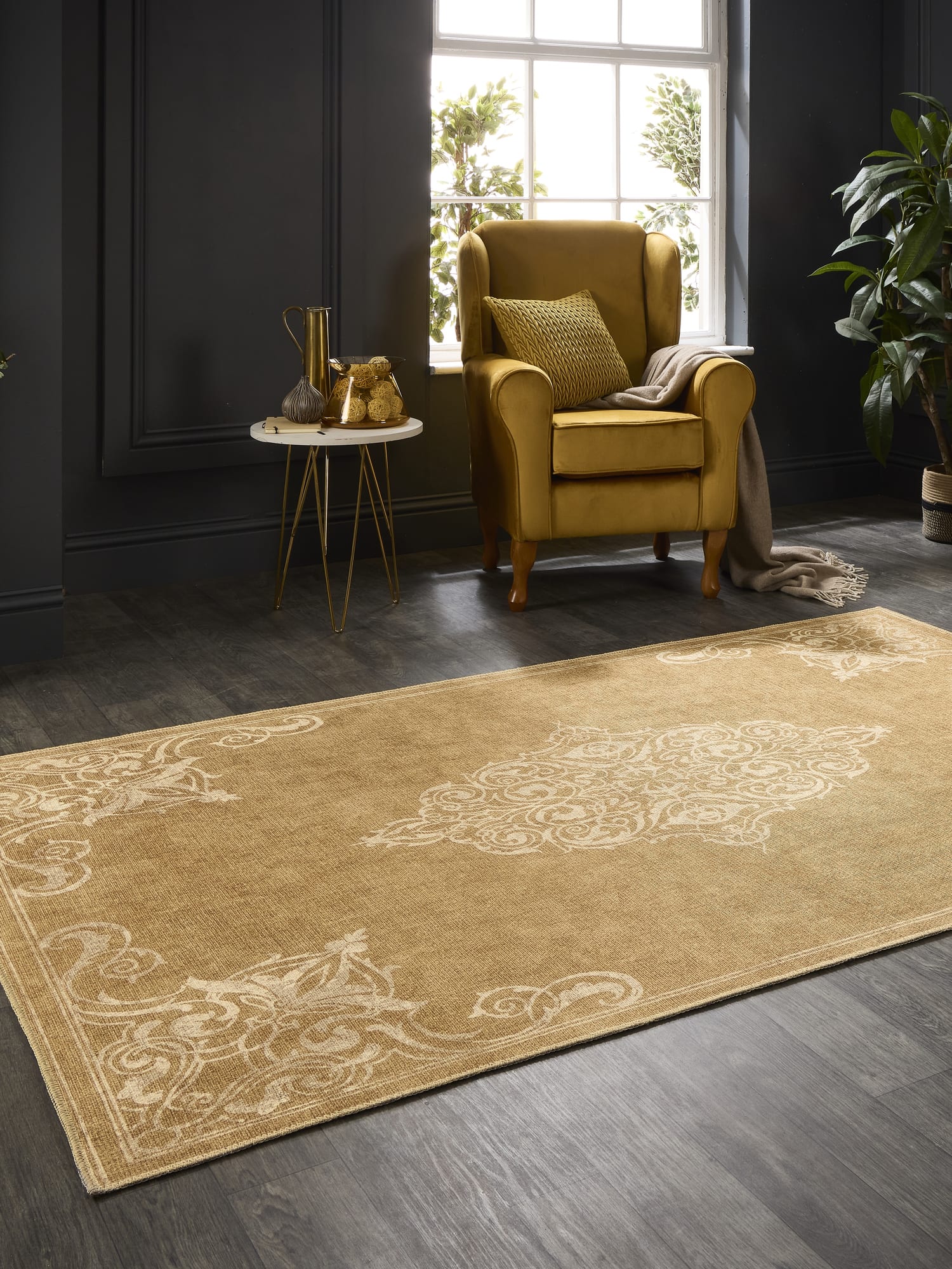 Sarabande rectangular rug in gold, at an angle beside an armchair in a dark-walled room.