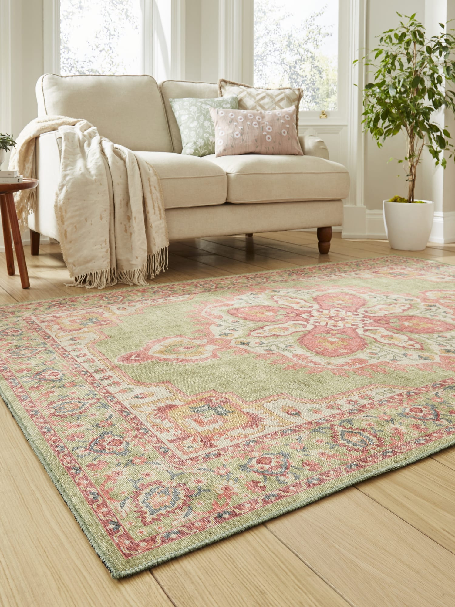 Zahra Washable rectangular rug in green and pink, at an angle beside a pale sofa.